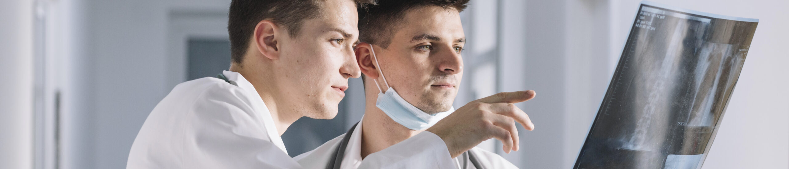 Dos profesionales de la salud, uno con cubrebocas en la barbilla, amos con bata analizando una radiografía en consultorio